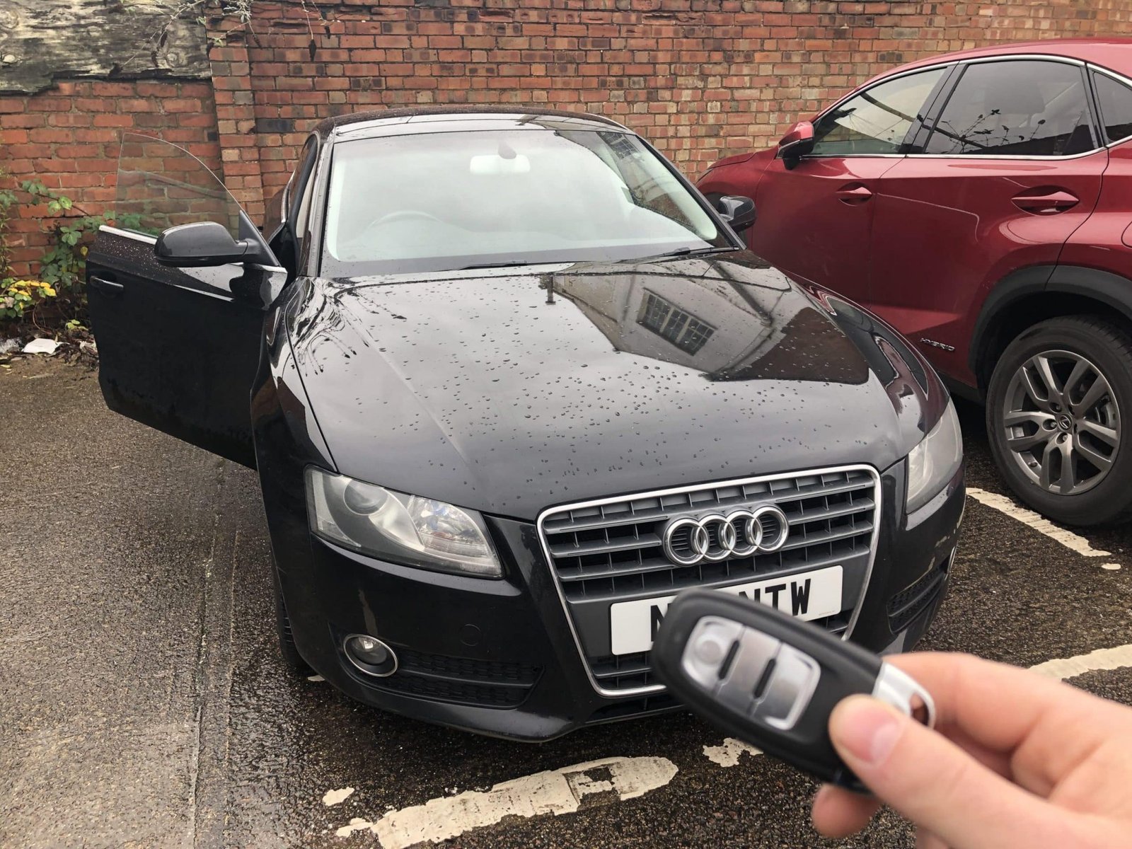 Audi key held in front of a black Audi with the driver door open